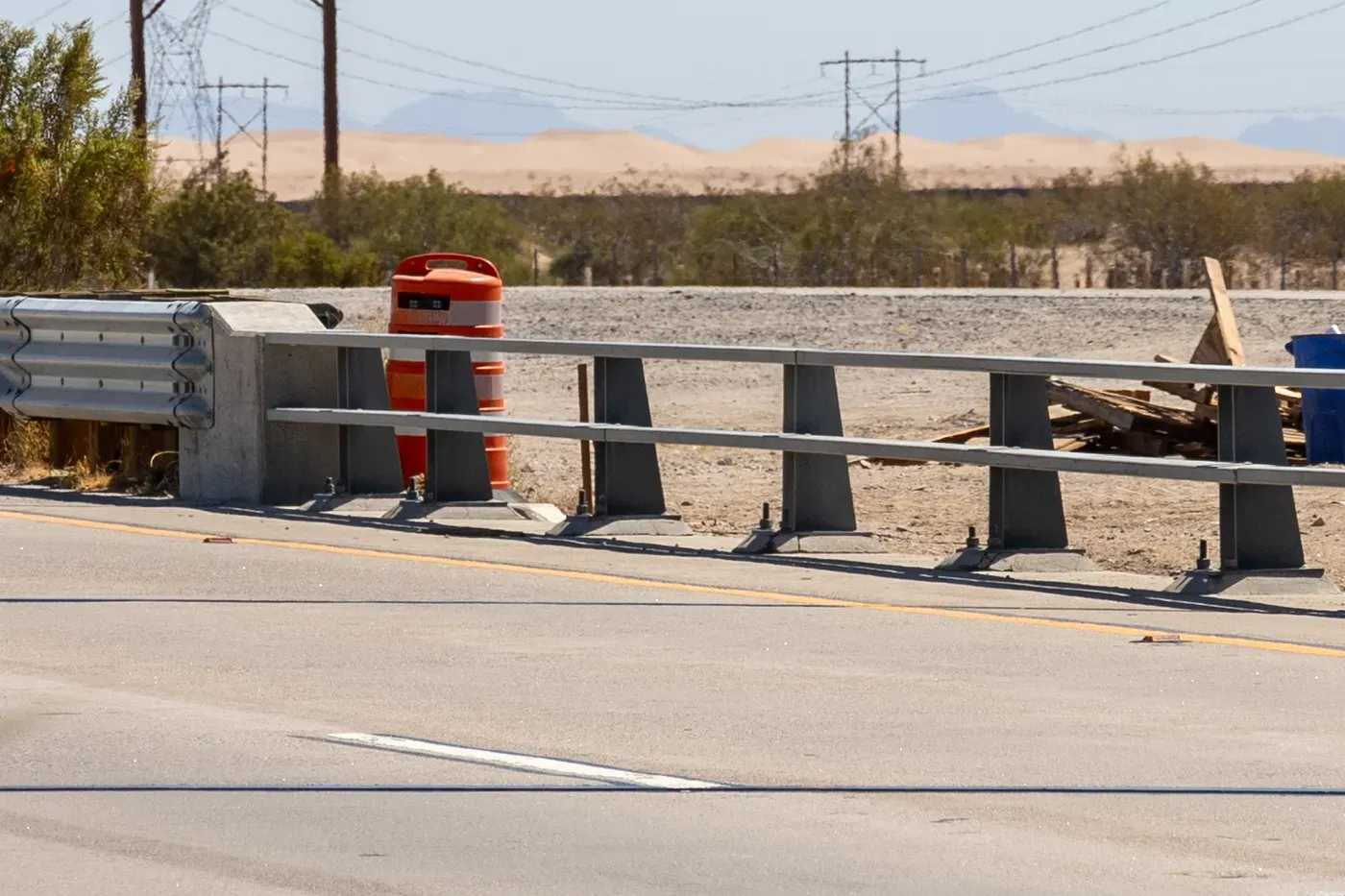 An orange traffic barrel at the roadside with a small camera unit mounted on top of it, beside a highway guardrail