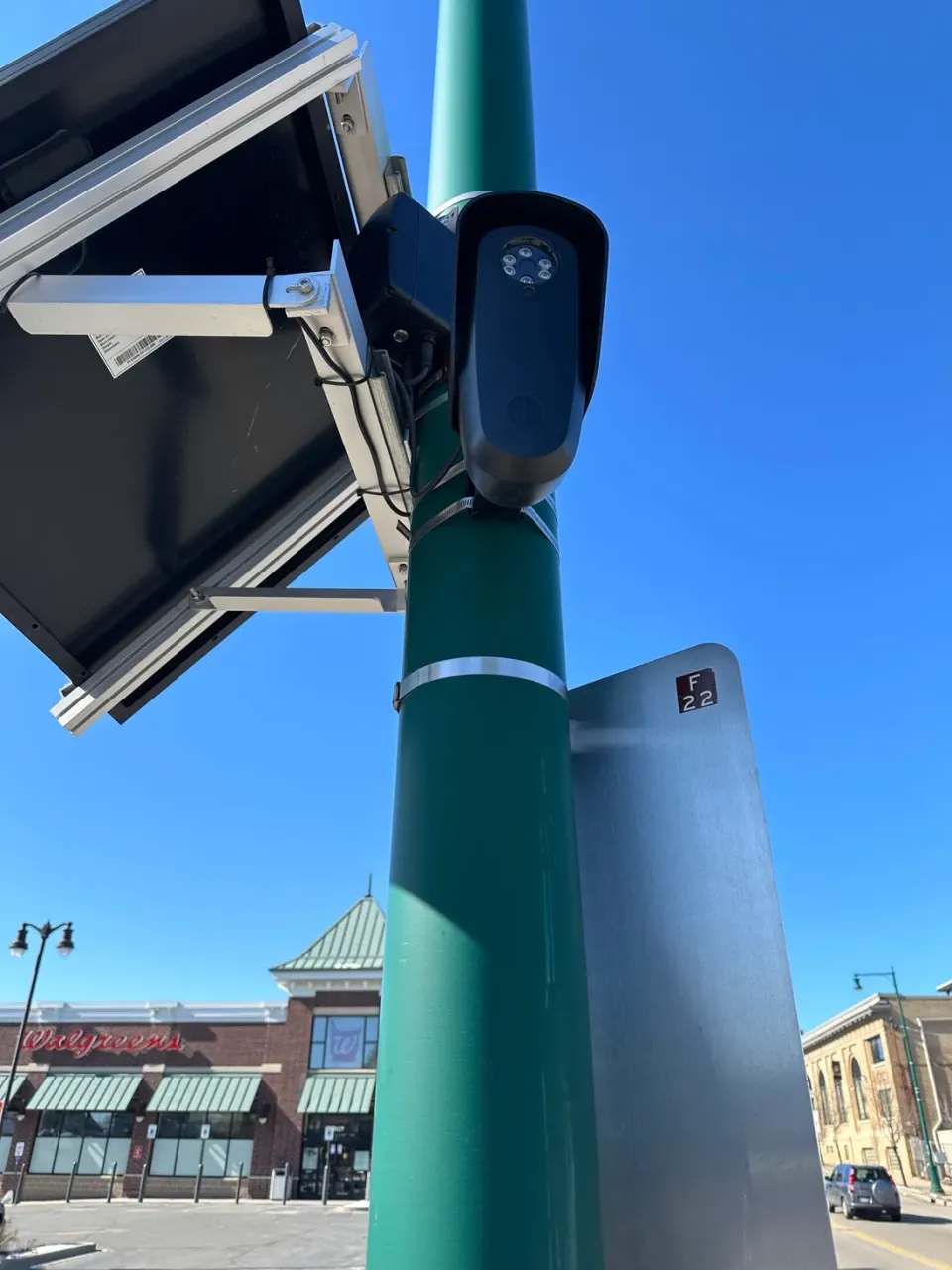 A license plate reader camera and solar panel mounted on a green pole on a small-town main street, with a pharmacy and brick storefronts behind it