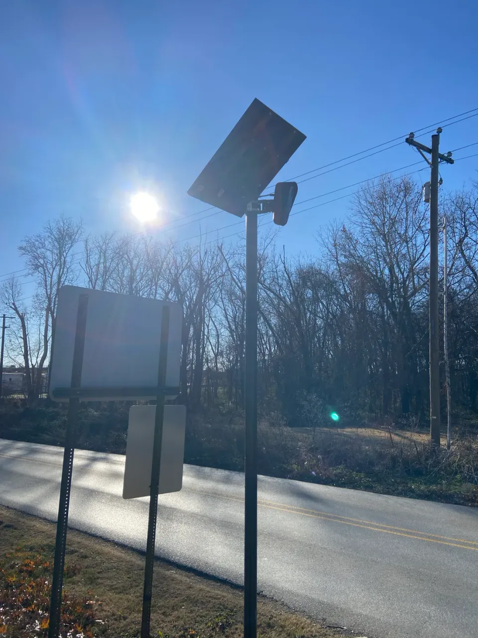 A license plate reader on a pole beside a rural two-lane road, backlit by low winter sun, with bare trees behind it