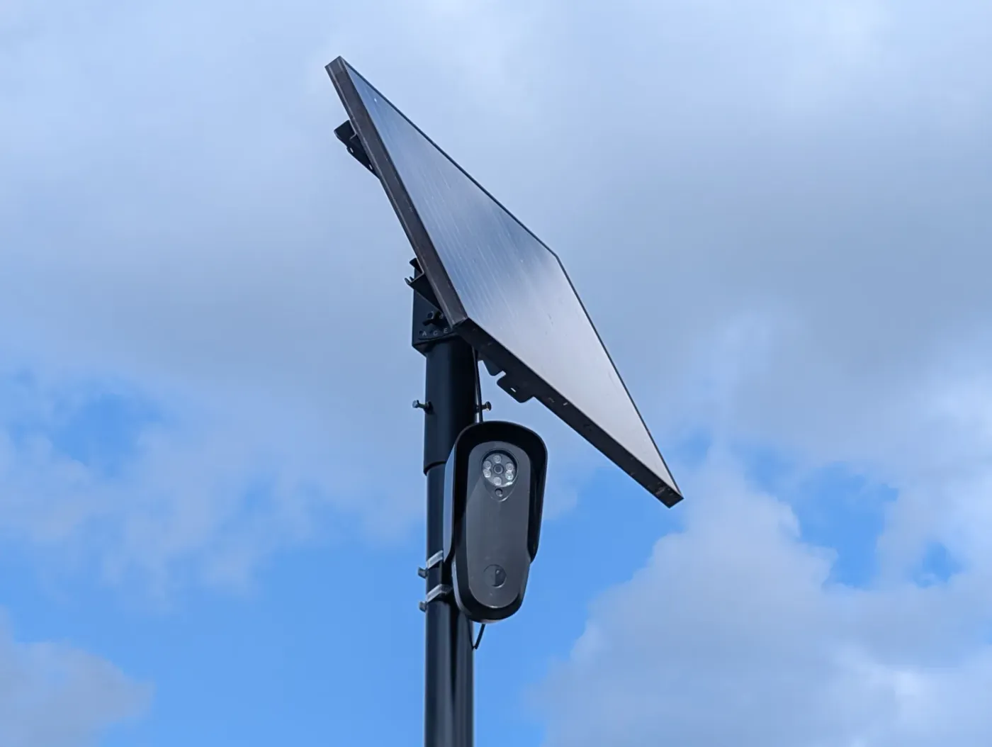 A license plate reader camera and its solar panel mounted on a black pole against a blue sky with clouds