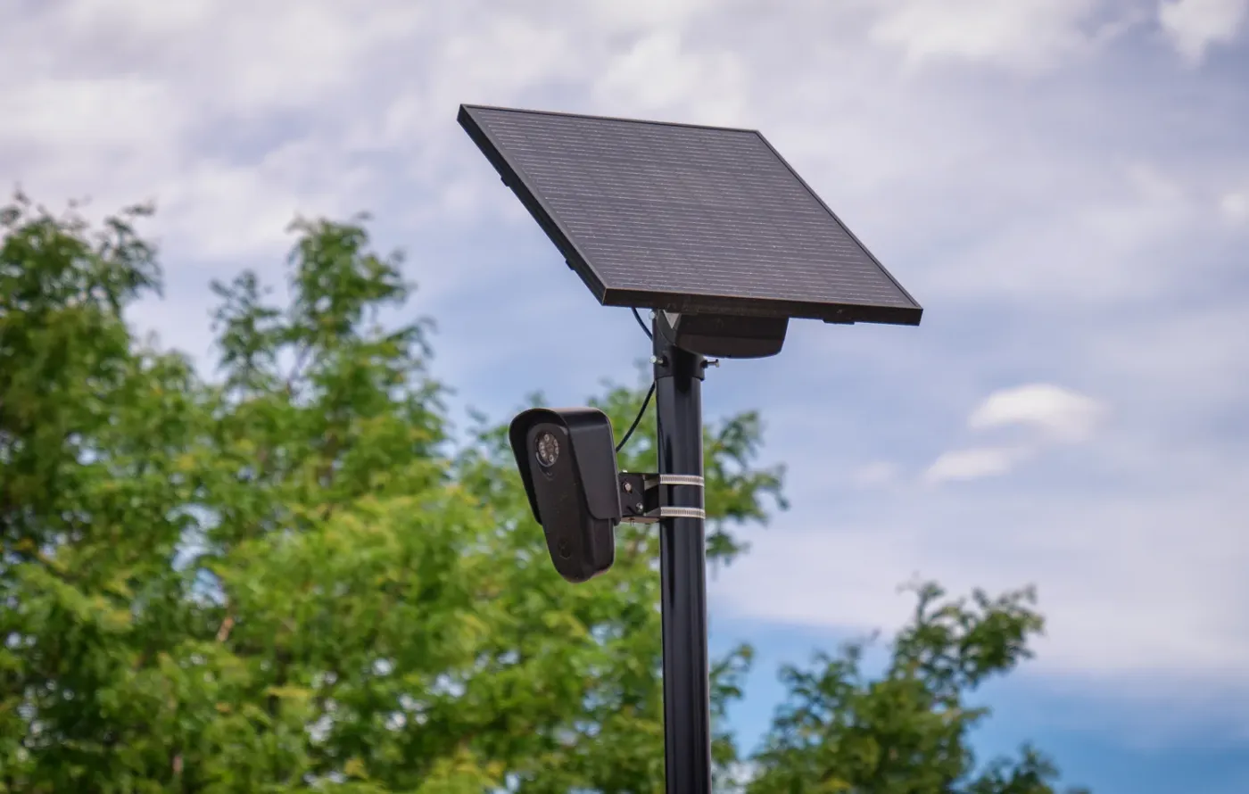 A close view of a license plate reader camera and its solar panel mounted on a black pole, with trees and sky behind